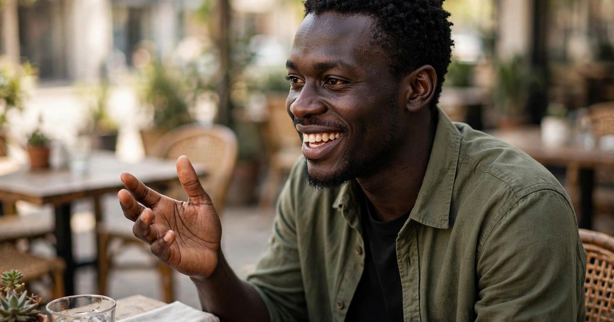 Illustration: AI-generated image of a fictional black West African man smiling warmly and gestures mid-conversation while seated alone at an outdoor cafe table.